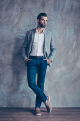 Full size portrait of stylish young bearded man standing on gray concrete background. He is in a suit, standing with crossed legs