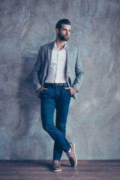 Full Size Portrait Of Stylish Young Bearded Man Standing On Gray Concrete Background. He Is In A Suit, Standing With Crossed Legs