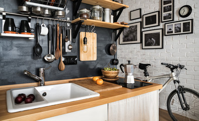 Design of modern home kitchen in the attic and rustic style. Black wall with shelves, trays, jars, mugs, sink. Against a wall with photos of a couple and a mountain bike.
