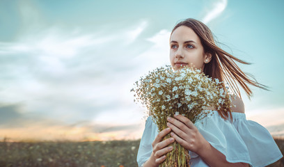 Beautiful girl is standing in a  field holding a bouquet of white flowers in her hands.