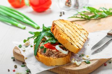 Fototapeta na wymiar Sandwich made of grilled toasts, tuna salad, tomato, onion and arugula on a white wooden table. Delicious healthy meal with ingredients.