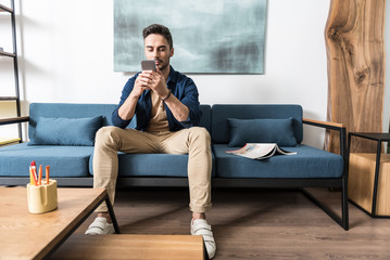 Obraz premium Concentrated youthful bearded guy using his smart phone in apartment