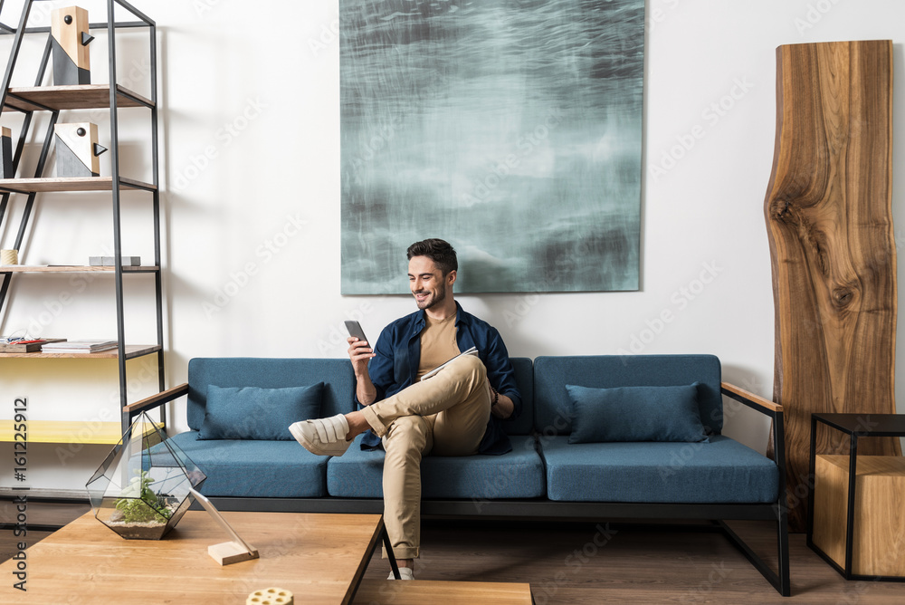 Wall mural happy youthful guy bearded resting with cellphone in living room