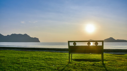 A lonely bench on green grass near the seashore at Ao Manao in Prachuap Khiri Khan province in southern of Thailand with beautiful blue sky, white cloud, morning golden sunlight and sea as background.
