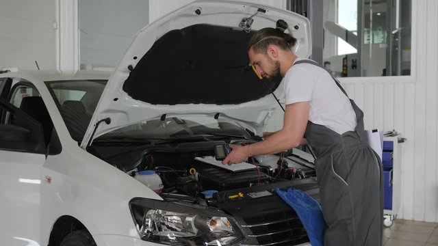 voltmeter in hands, auto mechanic measures battery voltage, mechanical data record service book, technical inspection machine, white car with an open hood, repair vehicle damage