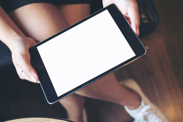 Mockup image of woman's hand holding black tablet pc with blank white screen on thigh with wooden floor background in modern cafe
