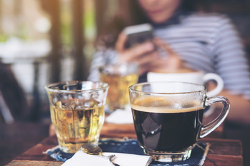 Closeup image of cups of hot coffee and tea on vintage wooden table in cafe with blur woman using smart phone in background