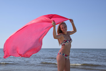 Blond girl in a bikini standing on the beach and holding a red silk handkerchief. Beautiful young woman in a colorful bikini on sea background