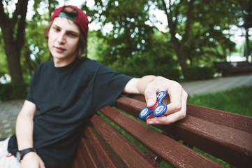 A long-haired hipster in a cap sits on a bench and spins a fidget-spinner