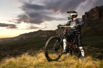Naklejka na ściany i meble The rider in full protection on a mountain bike stands and looks at the sunset on the background of the rocks