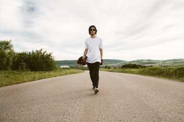 A guy with a longboard in his hands in a helmet and sunglasses is walking along an asphalt suburban road