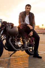 Handsome rider guy with beard and mustache in black leather biker jacket, boot and jeans sit on classic style cafe racer motorcycle. Bike custom made in vintage garage. Brutal fun urban lifestyle.