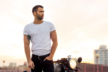 Sporty biker handsome rider male in white blank t-shirt walk away from classic style cafe racer motorbike at sunset. Vintage bike custom made in garage. Brutal urban lifestyle. Outdoor portrait. - 161269168