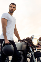 Sporty biker handsome rider male in white t-shirt hold black helmet near classic style cafe racer...