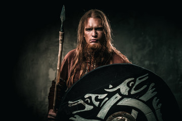 Serious viking with a spear in a traditional warrior clothes on a dark background.