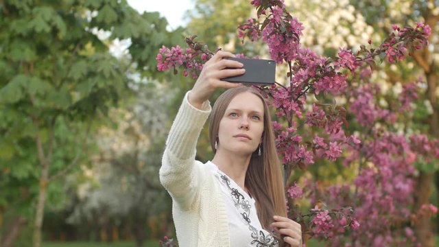Girl makes selfie in the garden. An attractive red-haired woman smiles making selfi using a mobile phone in a cherry orchard. The concept of using gadgets for a healthy lifestyle.