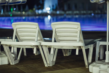 Sun beds by the pool at night. A row of empty armchairs for sunbathing and rest under the sun stand on the edge of the swimming pool. The territory is illuminated by colored lanterns.