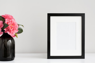 Mock up black photo frame on a white desktop next to a fresh bunch of pink camellia flowers in a...