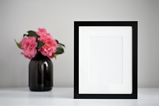 Mock up black photo frame on a white desktop next to a fresh bunch of pink camellia flowers in a black vase.