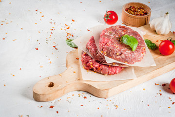 Fresh raw home-made minced beef steak burger with spices, tomatoes and basil, on a white stone concrete table, copy space
