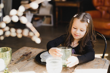 Pretty and cute little girl sitting near the table smiling. Adorable child in white lace dress. Childhood, happiness, joy, playing, celebration, holidays concept.
