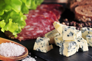 Roquefort slices on a dark board with a salt, salad, salami slices, peppercorns and bread