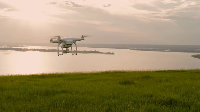 the white drone flying over the hill with green grass and is removed into the distance at sunset