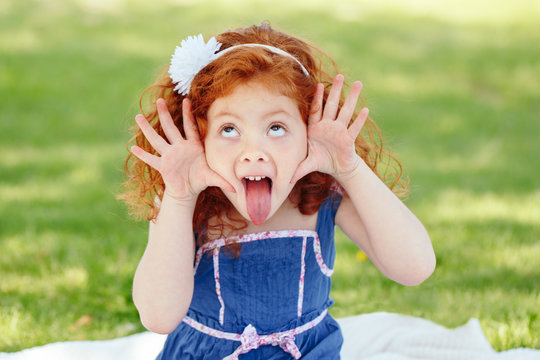 Portrait of cute adorable little red-haired Caucasian girl child in blue dress making funny silly faces showing tongue, in park outside, playing  crying screaming, having fun, lifestyle childhood