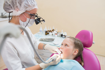 Young female dentist in loupe binoculars checked and curing teeth of child patient sitting in dental chair.