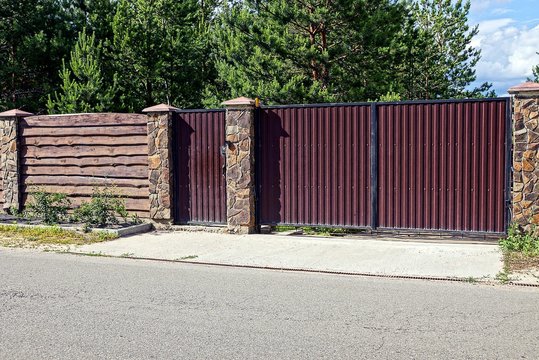 серо коричневые железные  ворота и деревянный забор перед асфальтированной дорогой 