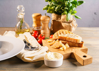 Antipasti set of toasts with pieces of parmesan on the dining-room board, decorated with cherry tomatoes, pot with basil leaves, bottle of olive oil, pepper and salt mills. Cutlery on a linen napkin.