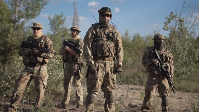 The commander in a military uniform and with weapon stand against the background of soldiers and looks in the camera