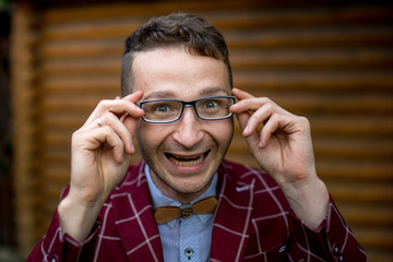 Portrait of stylish handsome young man with bristle standing outdoors. Man wearing jacket, glasses and shirt. Smiling man  in a red suit near a wood house