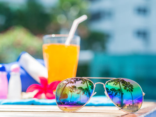 Red frangipani (plumeria) flowers, sungrasses, shampoo, lotion, hat, rolled up towels and Orange juice with a piece of lemon at the side of swimming pool. Vacation, beach, summer travel concept