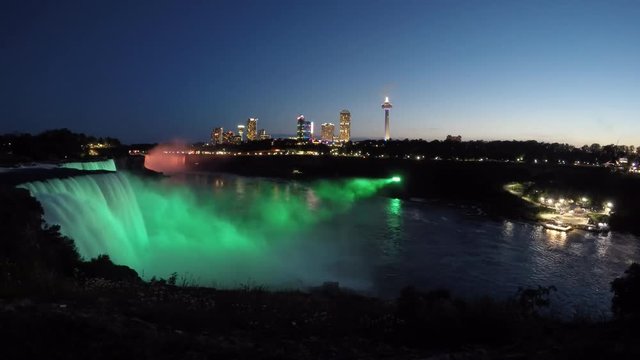 Niagara Falls colorful light illumination from New York State with Canadian skyline. Evening tour boat sailing into mist on Niagara River under fireworks. Time lapse