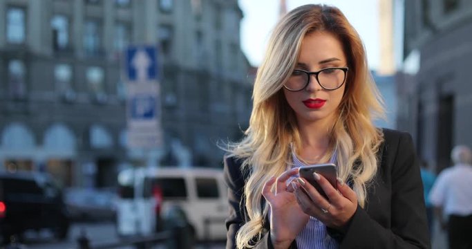 Close up view of a beautiful nerdy like serious businesswoman with red lipstick using her cellphone, she gets the message, and reacts happily to it, smiling brightly and texting back. Social networks.