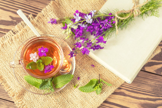 Glass cup of summer herbal tea with fresh mint and field larkspur. Bouquet of wild flowers on the notebook. Wooden table. Shallow depth of field.