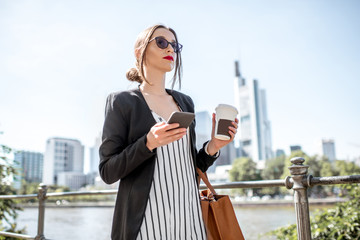 Young and elegant businesswoman walking with phone and coffee on the quay with skyscrapers in Frankfurt city