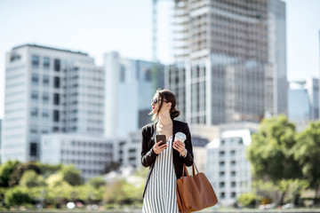 Young businesswoman walking with coffee and phone outdoors on the skyscrapers background in Frankfurt