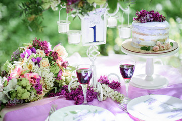 Table number one with bouquet of violet flowers and little cake on it stands in the garden