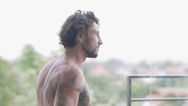 Handsome muscular man with tattoos on his bare back step out onto the open balcony