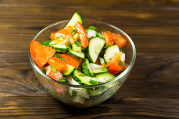 Fresh salad with tomato, cucumber, onion, parsley and dill in glass bowl on wooden table