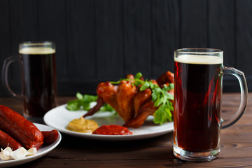 Two mugs of dark beer with crusty grilled chicken wings and various sausages.