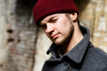 Bearded young man in red hat and grey coat poses before beige wall