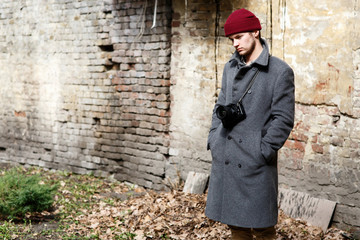 Serious young man with camera holds his hands in the pockets posing before ruined wall