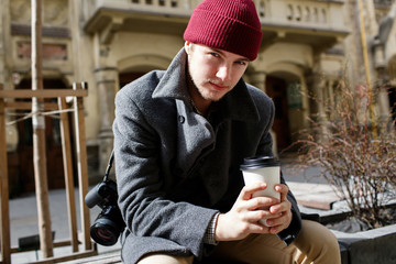 Young man in grey coat sits on bench with camera on his shoulder