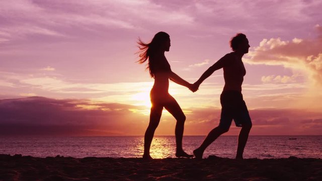 Romantic couple walking on beach holding hands at sunset with beautiful light and colors. Young fit couple on honeymoon enjoying travel vacation summer holidays on beach. SLOW MOTION RED EPIC.