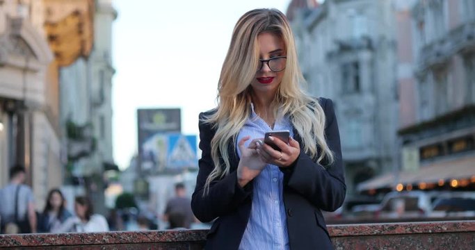 Pretty blonde woman in a business suit with red lips standing by the underground passage, and actively using her phone for texting, reacting happily, smiling brightly, texting back, stroking her hair.
