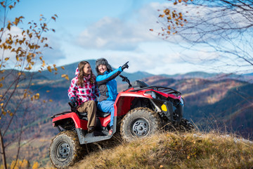 Fototapeta na wymiar Woman is showing something in distance to her friend in nature on the autumn sunny day. Two girlfriends in winter clothing on a red ATV at a hill on the background of mighty mountains and blue sky