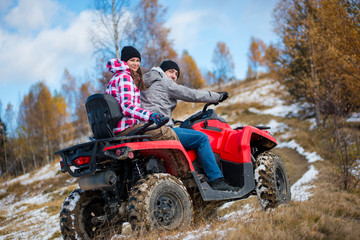 Fototapeta na wymiar Girl sitting behind man hugging him on red four-wheeler ATV and looking to the camera at snowy hill in the mountains. Beautiful landscape of nature in winter. Bottom view